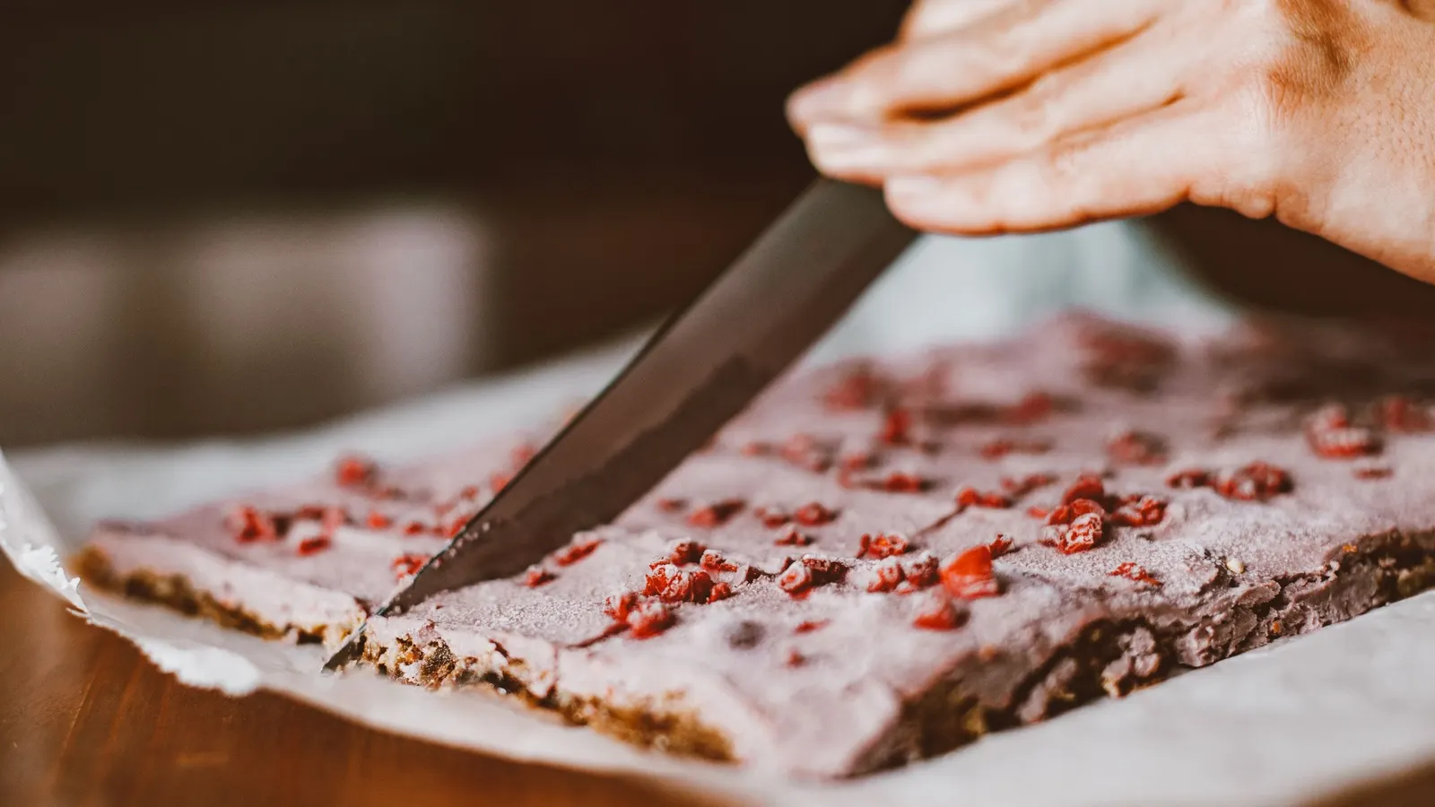 A pale fruit-set slab studded with bright red dried berry pieces being cut into bars with a long knife on a wooden bench
